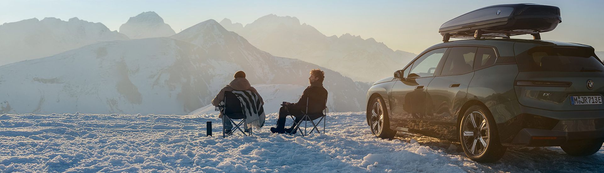 BMW iX mit Dachgepäckbox und zwei BMW Fahrern auf einer Skipiste
