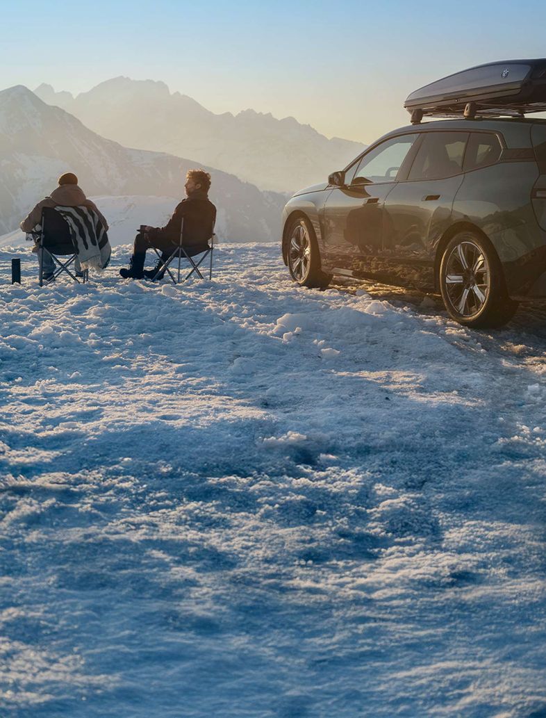 BMW iX mit Dachgepäckbox und zwei BMW Fahrern auf einer Skipiste